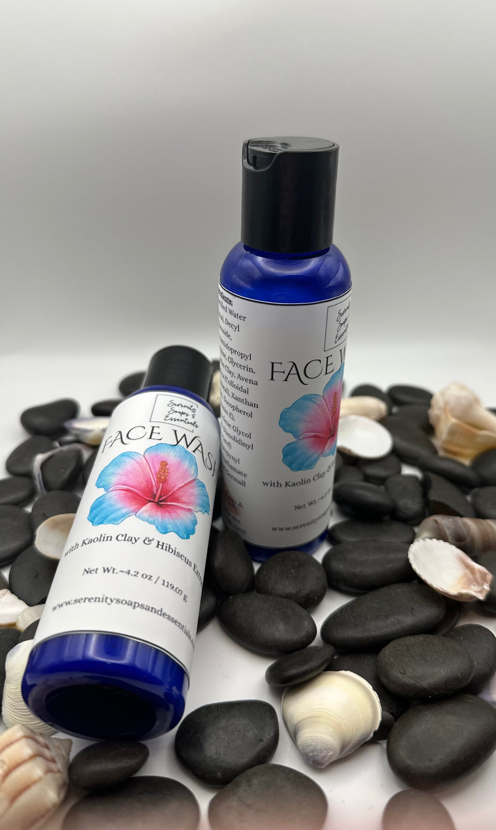 Two Face cleanser bottles with floral designs on a bed of pebbles and shells.