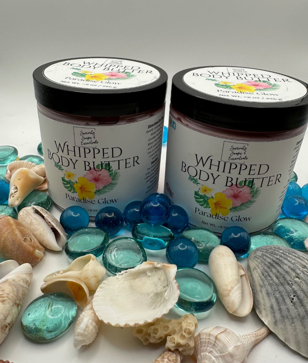 Two jars of 'Whipped Body Butter' with 'Paradise Glow' label surrounded by blue glass beads and seashells on a white background.