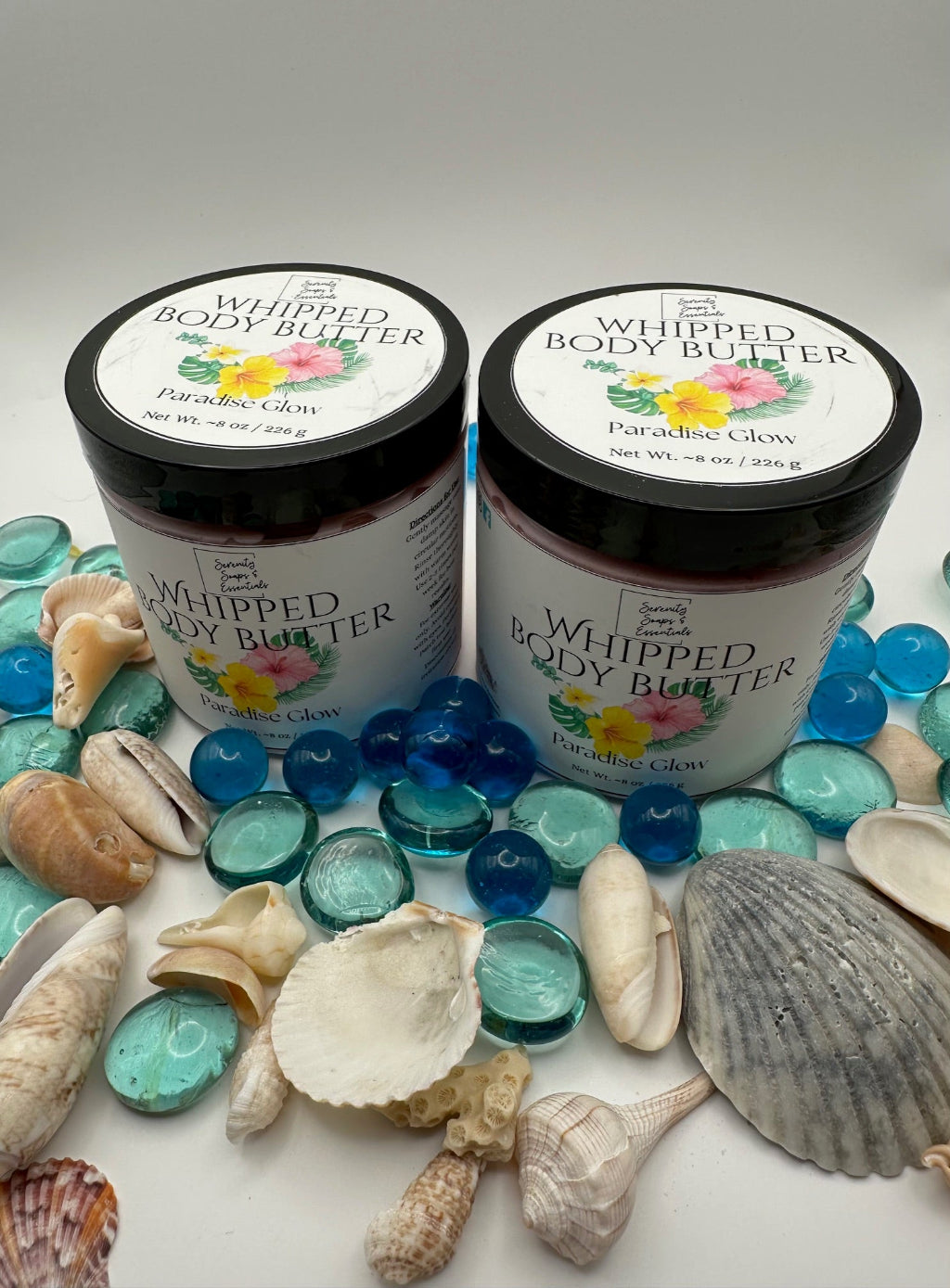 Two jars of whipped body butter with floral labels on a white surface with seashells and glass stones.