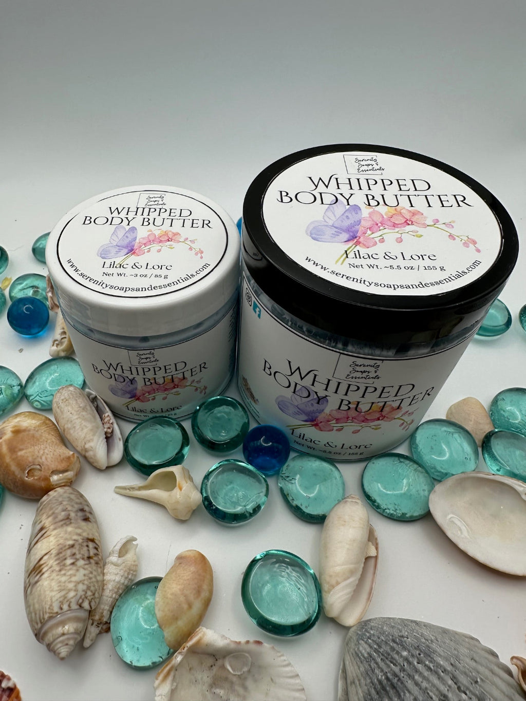 Two jars of whipped body butter with floral labels on a white surface with seashells and blue glass stones.
