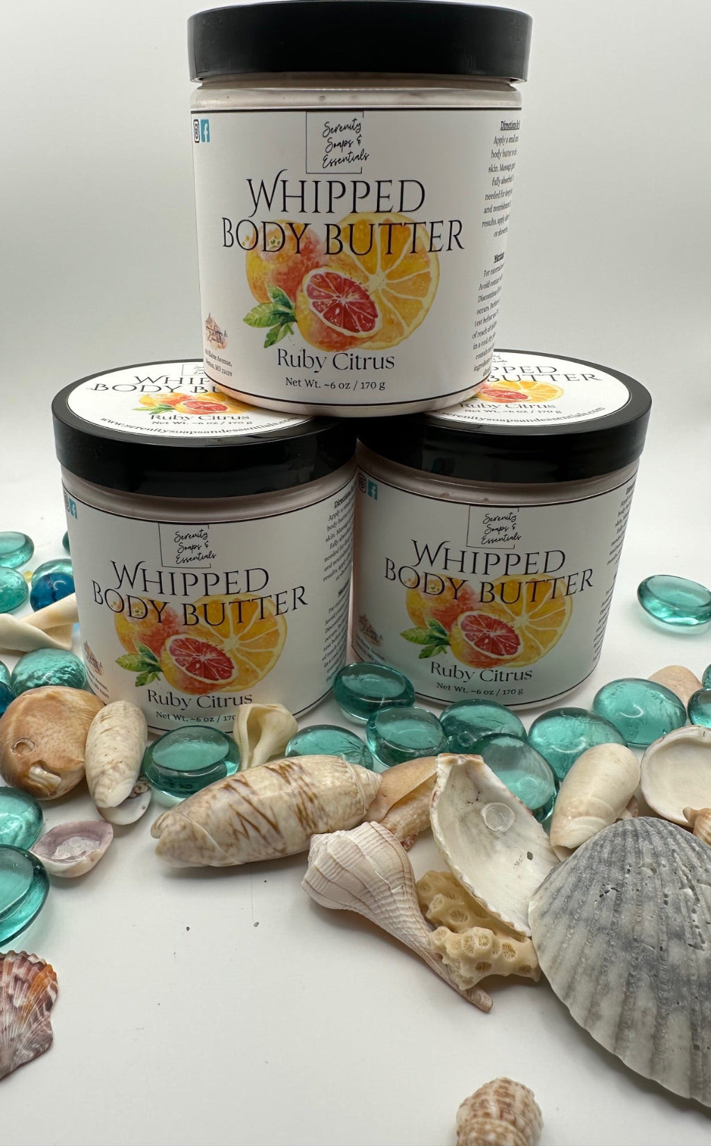 Three jars of Whipped Body Butter with Ruby Citrus flavor on a white surface with seashells and blue stones.