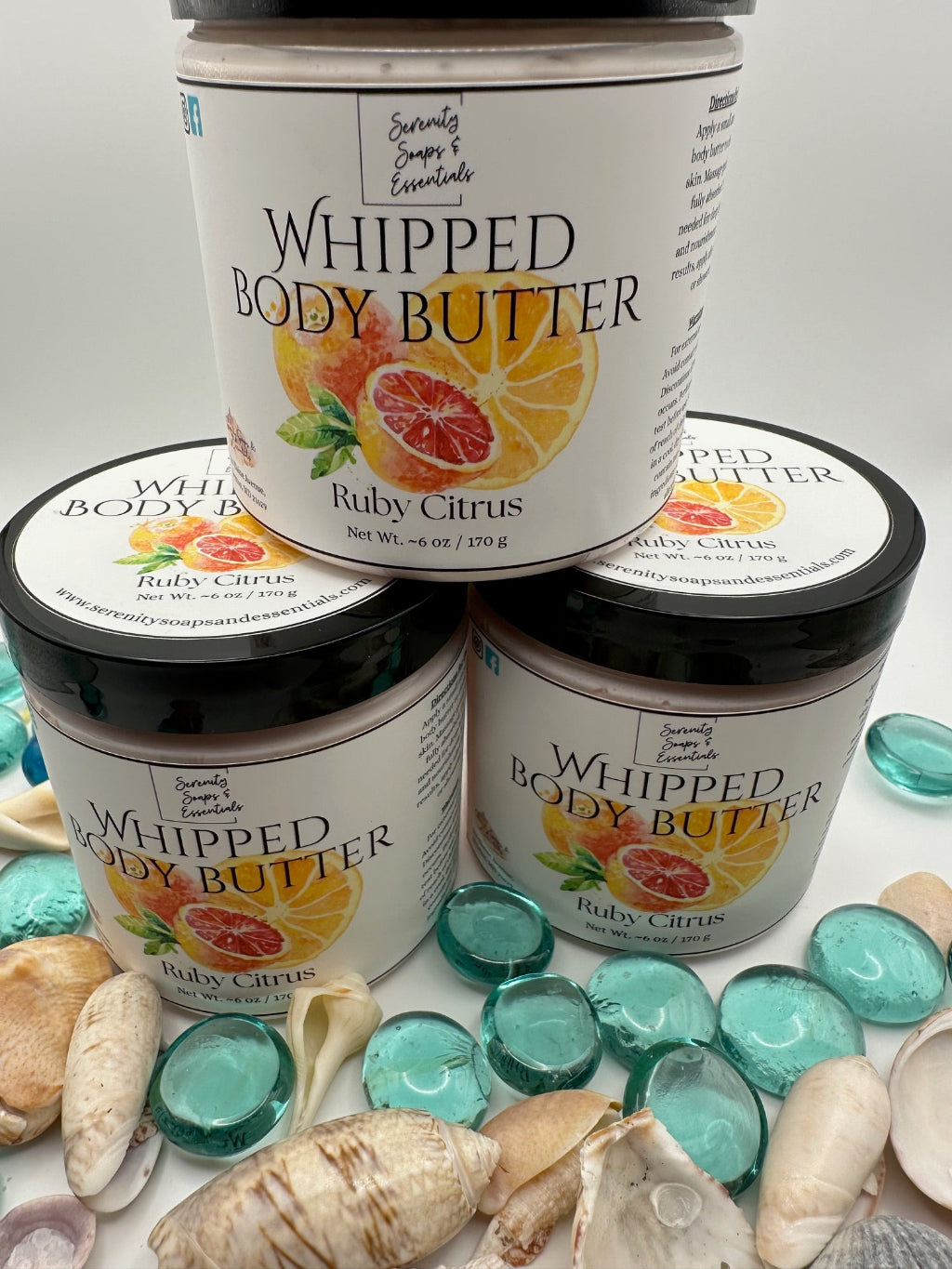 Three jars of 'Whipped Body Butter' with Ruby Citrus flavor on a surface with decorative stones and shells.