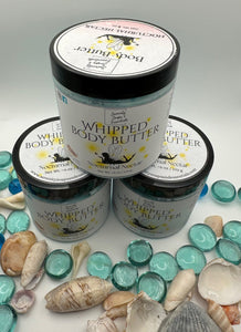 Three jars of whipped body butter with a label, surrounded by seashells and glass beads on a light gray background.