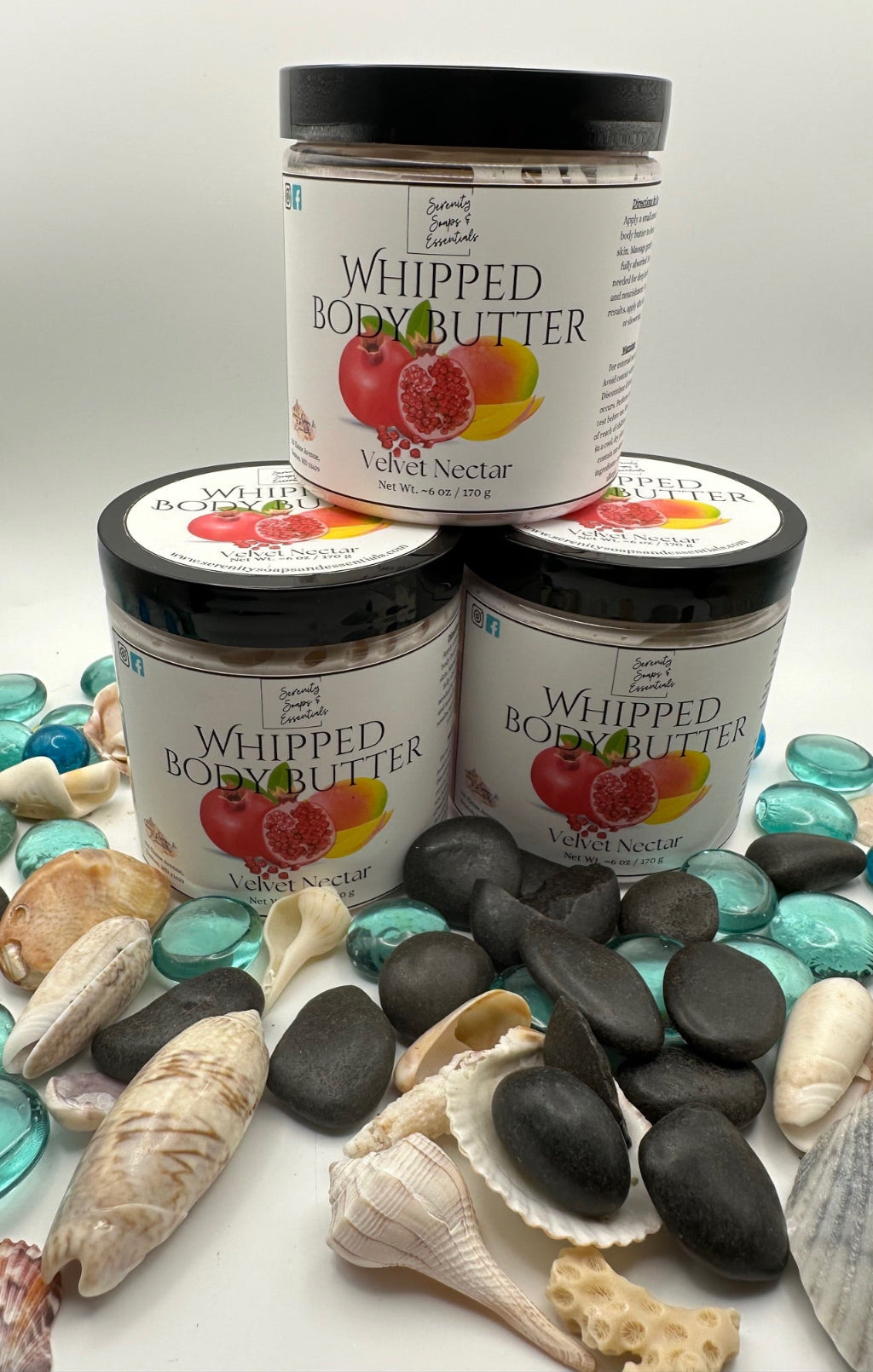 Three jars of Whipped Body Butter with a 'Velvet Nectar' flavor on a white background with pebbles and shells.