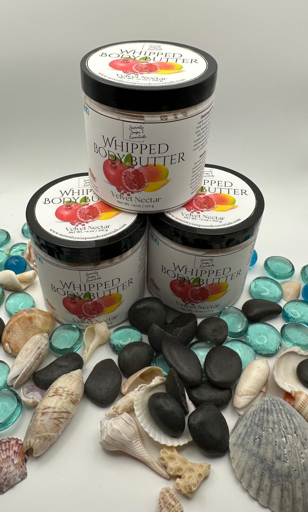 Jars of 'Whipped Body Butter' with beach-themed packaging on a white surface with seashells and stones.